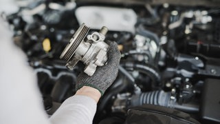 Zu sehen ist die Hand einer Person, die vor einem Auto mit geöffneter Motorhaube steht und ein Autoersatzteil hält.