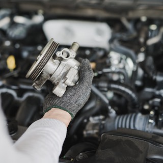 Zu sehen ist die Hand einer Person, die vor einem Auto mit geöffneter Motorhaube steht und ein Autoersatzteil hält.