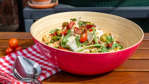 Mediterraner Nudelsalat ist in einer roten Salatschüssel angerichtet, daneben liege Salatbesteck und rote Cocktailtomaten.