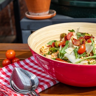 Mediterraner Nudelsalat ist in einer roten Salatschüssel angerichtet, daneben liege Salatbesteck und rote Cocktailtomaten.