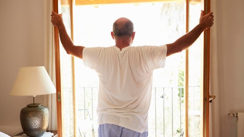 Ein Mann steht mit ausgebreiteten Armen an einem geöffneten Fenster im Schlafzimmer und schaut hinaus. Bei hohen Temperaturen im Sommer ist eine regelmäßige Lüftung gegen die Hitze wichtig.