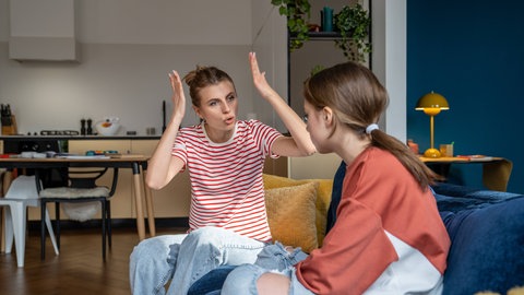 Verärgerte Mutter gestikuliert schreiend zu ihrer Teenager-Tochter, die zu Hause auf der Couch sitzt. Perfektionismus bei jungen Erwachsenen und Studierenden ist nicht nur eine Frage der Erziehung: Neben dem Elternhaus beeinflussen vor allem gesellschaftliche Werte, wie stark junge Menschen unter dem Druck leiden, fehlerlos sein zu müssen.