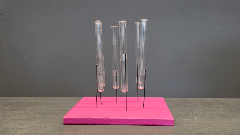 Löcher in die mit Abtönfarbe colorierte Holzplatte bohren und die vorbereiteten Reagenzgläser mit dem Draht einstecken. Vor dem einstecken Heißkleber anbringen. 