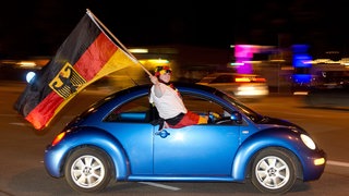 Junger Mann mit riesiger Deutschlandflagge sitzt bei voller Fahrt auf Autofenster von blauem Kleinwagen. Was ist beim Autokorso erlaubt? Darf ich Zimmer an Fußballfans untervermieten?