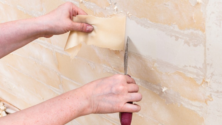 Eine Person zieht die Tapete mit Hilfe einer Spachtel von der Wand