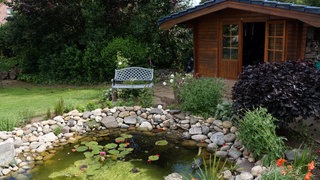 Neben dem runden Gartenteich mit Seerosen und Algen steht eine Bank auf dem grünen Rasen. Daneben ist eine hübsche Gartenhütte aus Holz mit offenen Türen zu sehen.