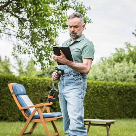 Ein Mann steht nachdenklich mit einem Tablet in der einen und dem Wassersprenkler in der anderen Hand auf dem Rasen im Garten.