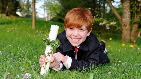 Ein Junge liegt am Tag seiner Erstkommunion auf einer Wiese und hält eine weiße Kerze in der Hand.