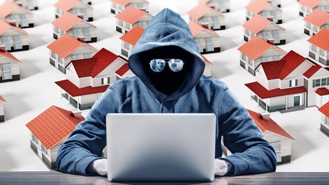 Symbolbild für Internetbetrüger bei Wohungsanzeigen: Eine vermummte, dunkle Gestalt mit Spiegelbrille sitzt vor einem Laptop. Im Hintergrund eine computergenerierte Landschaft mit Häusern.