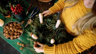 Eine junge Frau dekoriert einen Adventskranz mit Kerzen und kleinen Zapfen.