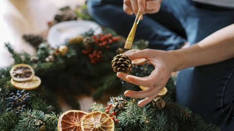 Eine Frau trägt mit einem Pinsel goldene Farbe auf einen Tannenzapfen für einen Adventskranz auf.