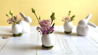 Osterdeko 2026: Eine Vase aus einem Hühnerei mit hübschen Blüten darin steht als Deko an Ostern auf dem Tisch.