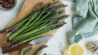 Grüner Spargel – Anbau im Garten: Frischer Grüner Spargel liegt auf einem Schneidebrett. Daneben bunte Pfefferkörner, ein Geschirrtuch, eine halbierte Zitrone und eine Knoblauchknolle.
