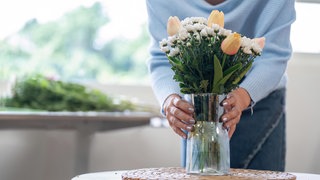Eine Frau hält einen Blumenstrauß in einer Vase und trägt ein blaues Oberteil. 