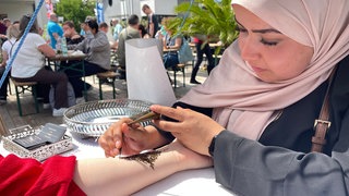 Eine Frau macht einer Besucherin ein Henna-Tattoo auf den Arm im afrikanischen Dorf auf dem Maimarkt in Mannheim. 