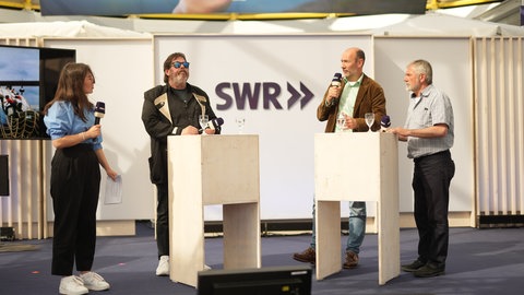 Frau interviewed drei Gäste auf der Bühne im Gläsernen Studio auf dem Gelände des Mannheimer Maimarktes. 