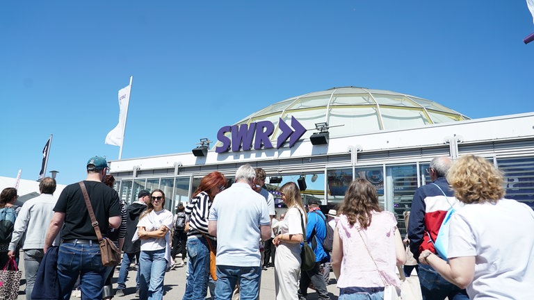 Besucher auf dem Mannheimer Maimarkt vor dem Gläsernen Studio des SWR bei sommerlichem Wetter.