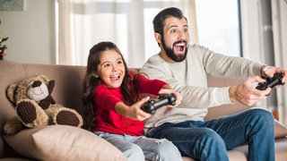 Ein Vater und seine Tochter sitzen auf der Couch und zocken gemeinsam.