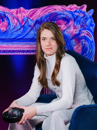 Host Sissy Hertneck sitzt mit Kopfhörer vor einem großen Bilderrahmen auf dunklem Hintergrund