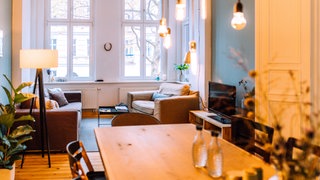 Blick in ein gemütliches Wohnzimmer: Am Fenster stehen zwei Sessel, von der Decke hängen Leuchten über einem großen Esstisch aus Holz. ARD-Mitmachaktion #besserwohnen