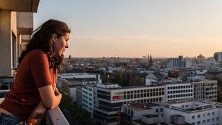 Junge Frau lehnt sich auf Balkon und blickt bei Sonnenuntergang auf die Skyline von Berlin. 