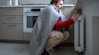 Ein in eine Decke eingewickelter Mann versucht sich an der Heizung zu. 