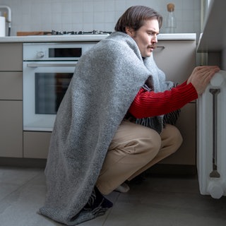 Ein in eine Decke eingewickelter Mann versucht sich an der Heizung zu. 