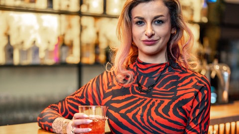 Eine Frau mit langen blonden Haaren, in einem engen roten Kleid, sitzt an der Bar. 