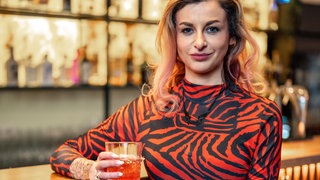 Eine Frau mit langen blonden Haaren, in einem engen roten Kleid, sitzt an der Bar. 