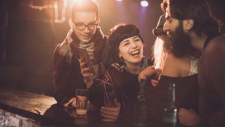 Eine dunkelhaarige Frau steht an der Bar und lacht. Um sie herum stehen andere Menschen. Sarah probiert es aus, und geht raus. Bild: I-StockSouth_agency