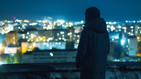 Eine Person alleine im Dunkeln, Lichter einer Stadt im Hintergrund. Bild: iStockEvgeny Gostuhin