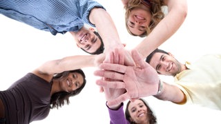Mehrere jungen Menschen legen die Hände übereinander und lachen. Freunde sind wichtig, aber manchmal auch kompliziert. Mehr unter EXIT EINSAMKEIT