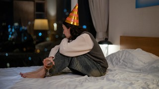 Eine Frau mit Partyhut sitzt alleine auf einem Bett, draußen ist es dunkel. Ein Tag, der immer weh tut – obwohl er eigentlich schön sein sollte. Sarah hat Geburtstag.  