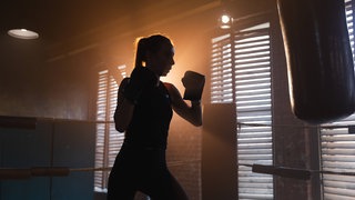 Boxerin steht vor Boxsack, die Sonne strahlt von hinten in den dunklen Raum hinein