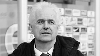 Eugen Strigel steht vor einer Pressewand und trägt eine Krawatte. Das Bild ist schwarz-weiß gefärbt.