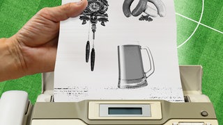 Ein altes Faxgerät, aus dem ein Blatt Papier kommt mit kleinen Bildern darauf: Kukuksuhr, Würste und ein Bierkrug. Eine Hand nimmt das Blatt aus dem Fax und im Hintergrund ein Fußballfeld.