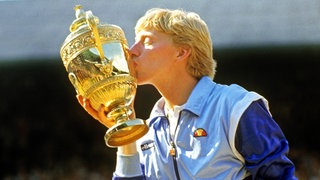 Boris Becker trägt eine blaue Jacke und küsst den Siegerpokal von Wimbledon