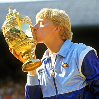 Boris Becker trägt eine blaue Jacke und küsst den Siegerpokal von Wimbledon