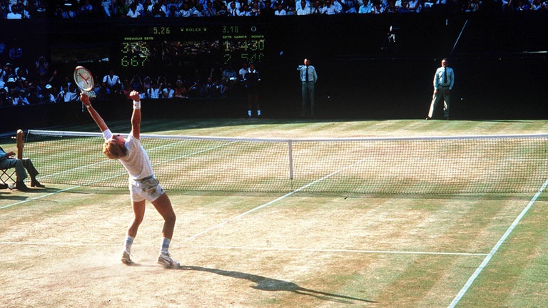 Boris Becker trägt ein weißes Tennis-Outift und reißt auf dem grünen Tennisrasen die Hände in die Luft, nach dem er den Matchball verwandelt hat.