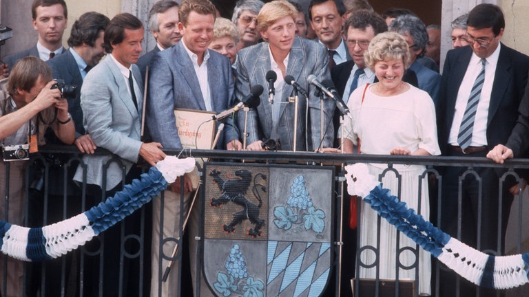 Boris Becker steht hinter einem Sprecherpult auf dem Rathausbalkon von Leimen. Um ihn herum stehen der Bürgermeister und viele andere Personen.