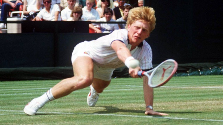 Boris Becker trägt ein weißes Tennis-Outifit und hechtet mit seinem Schläger nach einem Ball