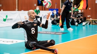 Sitzvolleyballer Martin Vogel sitzt im schwarzen Trikot der Nationalmannschaft auf dem Spielfeld, den grün-rot-weißen Volleyball hat er mit der linken Hand angeworfen, mit der rechten Hand hat er ausgeholt, um beim Qualifikationsturnier für die Paralympics 2021 in Tokio aufzuschlagen.