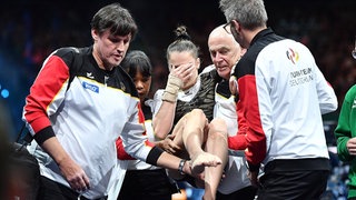 Helen Kevric verdeckt sich das Gesicht, während die Turnerin von Betreuern von der Matte getragen wird.