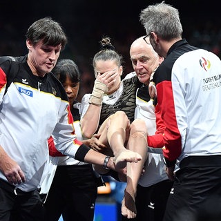 Helen Kevric verdeckt sich das Gesicht, während die Turnerin von Betreuern von der Matte getragen wird.