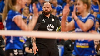 Konstantin Bitter, Trainer von Allianz MTV Stuttgart, steht an der Seitenlinie und betrachtet ein Spiel seiner Volleyballerinnen. Unscharf im Vordergrund: Zwei Spielerinnen in blaunen Trikots klatschen sich ab und rahmen den Trainer damit ein.