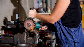 Eine Frau arbeitet im Beruf als Metallbauerin in einem Betrieb des Metallbau. Mit dem Winkelschleifer schneidet Sie ein Stück Metall auf Mass.