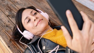 Frau mit Kopfhörern und Smartphone liegt auf dem Holzboden. Radio und Podcast hören wann und wo ihr wollt.