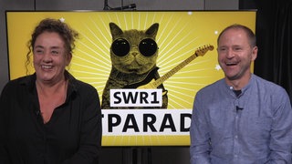 SWR1 Moderatorin Annett Lorisz und Patrick Neelmeier sitzen nebeneinander und lachen in die Kamera