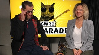 Die SWR1 Moderatoren Stefanie Anhalt und Jochen Stöckle sitzen auf Hockern. Stefanie lächelt in die Kamera, Jochen rückt seine Sonnenbrille zurecht.