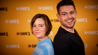 Die SWR1 Moderatoren Cora Klausnitzer und Matthias Sziedat stehen mit dem Rücken aneinander und lächeln in die Kamera.